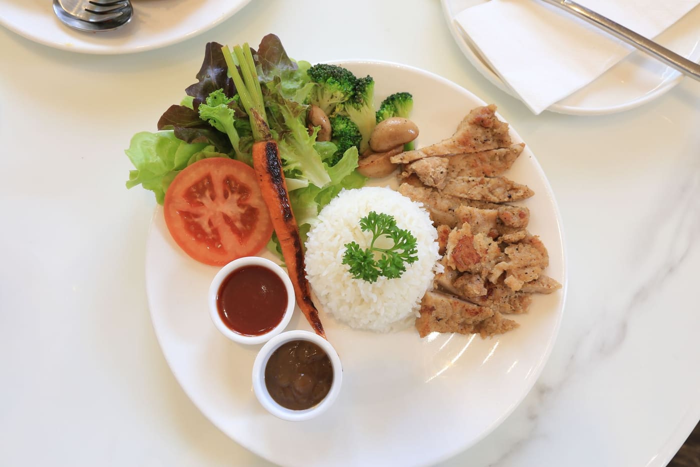 A white plate of grilled chicken, rice, salad vegetables and two small sauce dishes