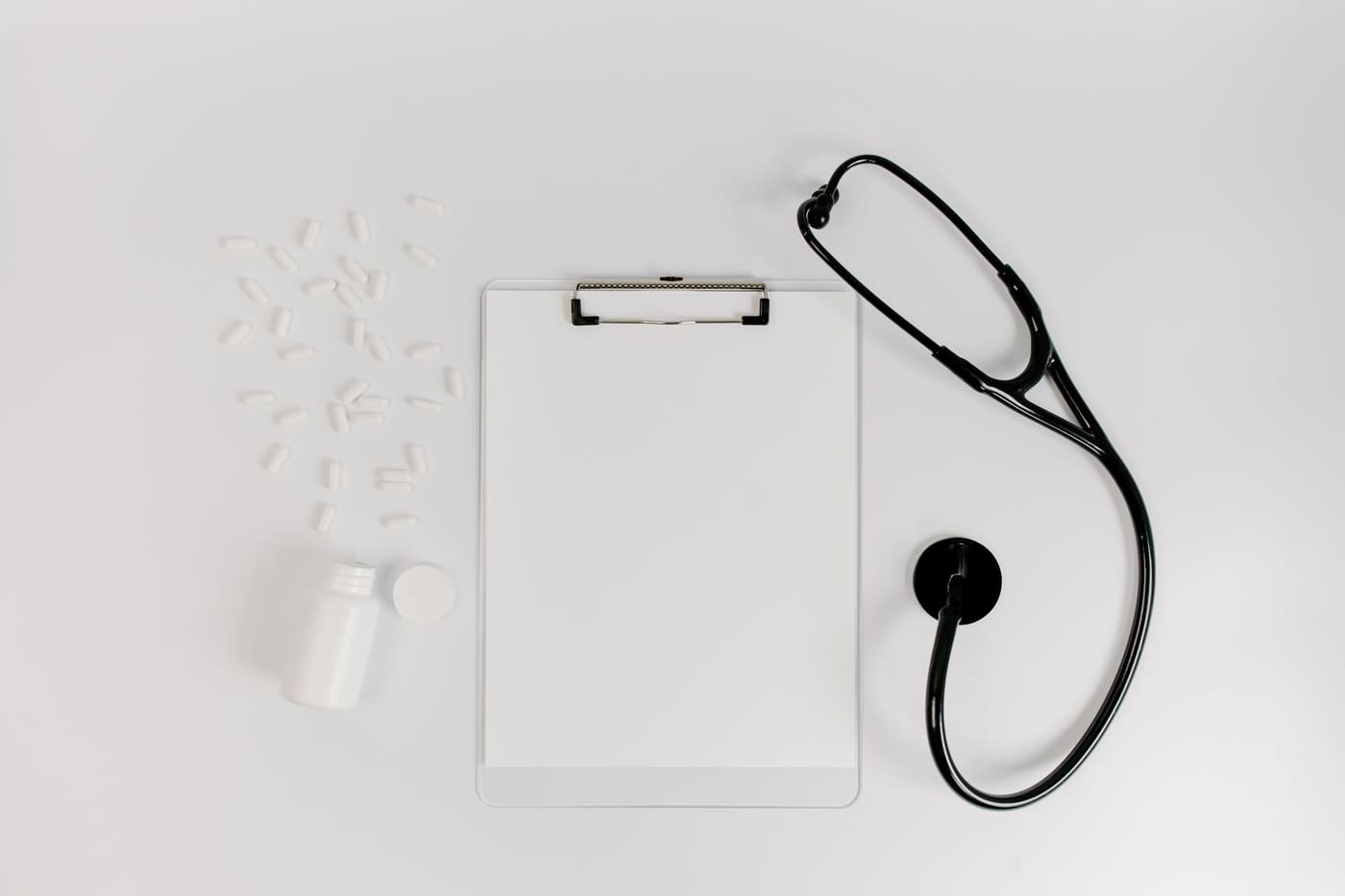 A clipboard with blank paper, a stethoscope and pill bottle laid flat on a white surface